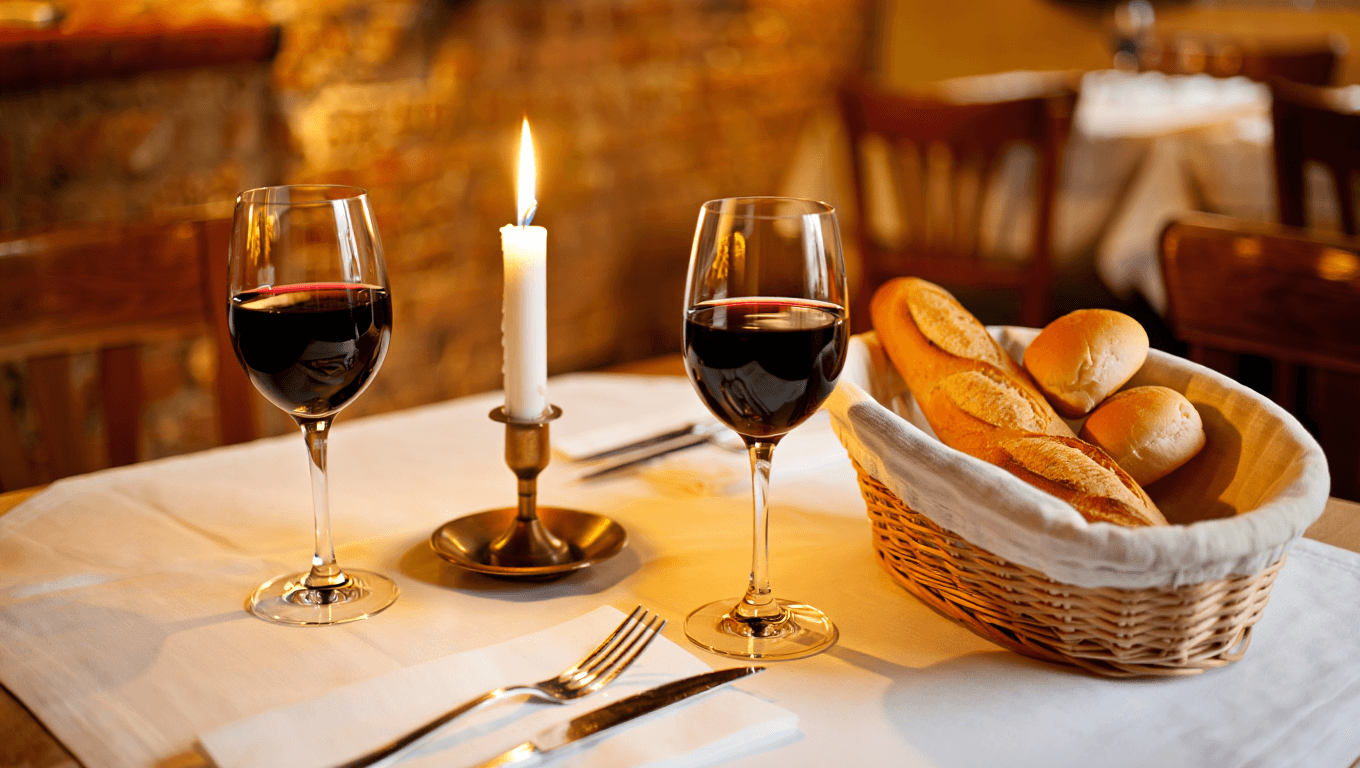 Cozy French bistro table set with white linen, candlelight, wine glasses, and a rustic bread basket in warm amber light